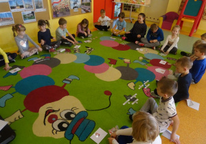 Grupa dzieci siedzi na dywanie z rozłożonymi obrazkami, wybierają z pośród różnych herb Warszawy.